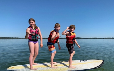 Three kids in lifejackets balance on waterboard